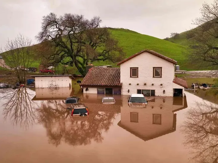 Floodwater surrounding a two-story house and partly submerged parked cars after a storm